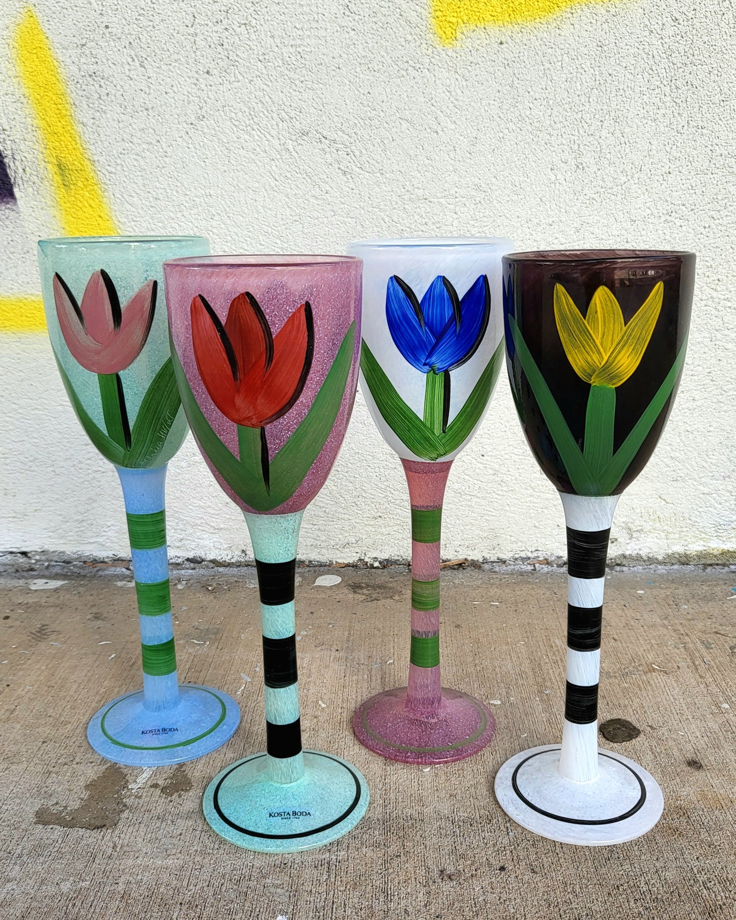 Set of four hand-painted Kosta Boda glass goblets by Ulrica Hydman-Vallien, each featuring a tulip design in different colours with striped stems, displayed on a wooden table