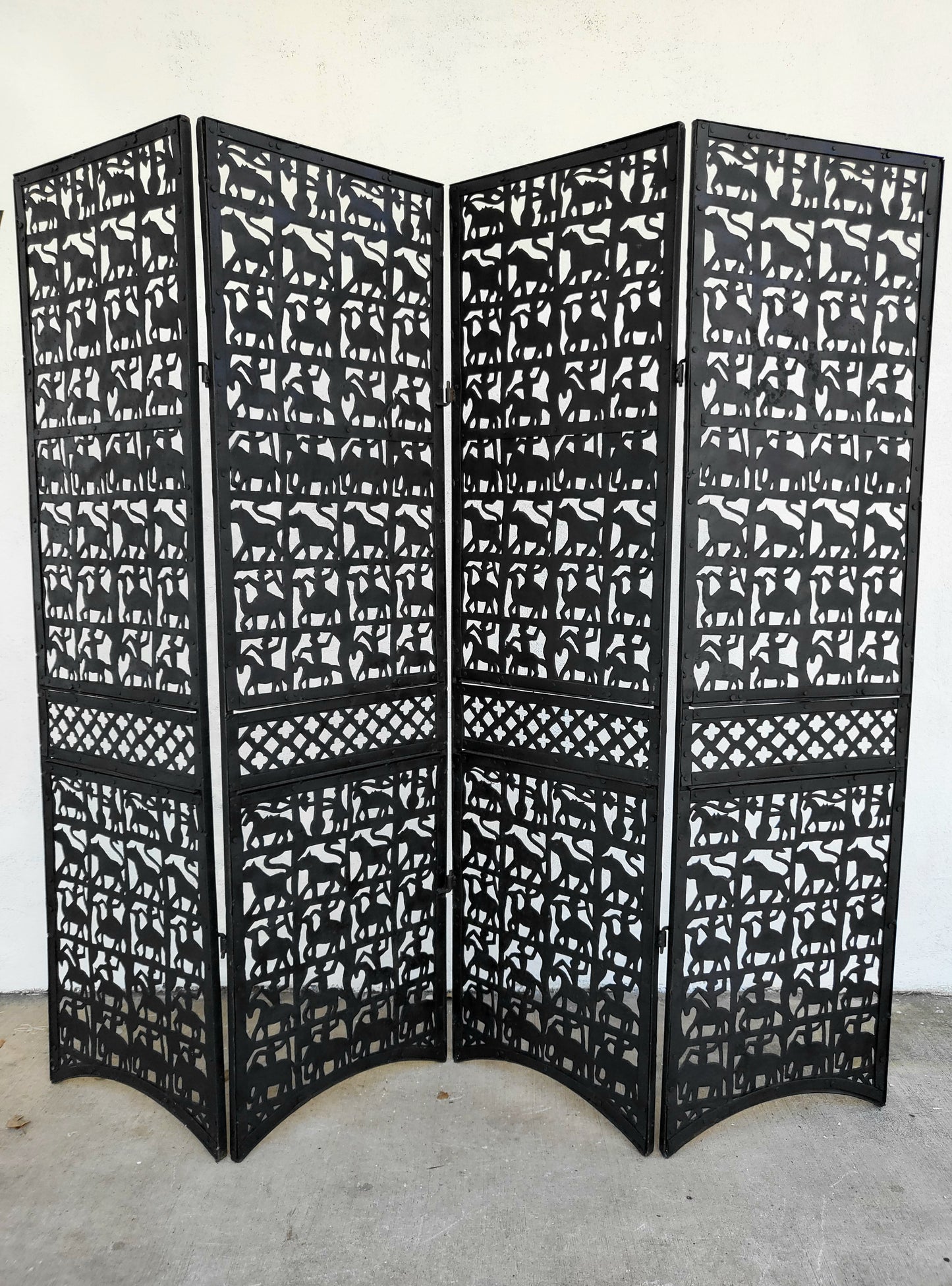 Four-panel black metal folding room divider with hand-cut animal silhouettes and lattice detailing, standing outdoors against a white wall.