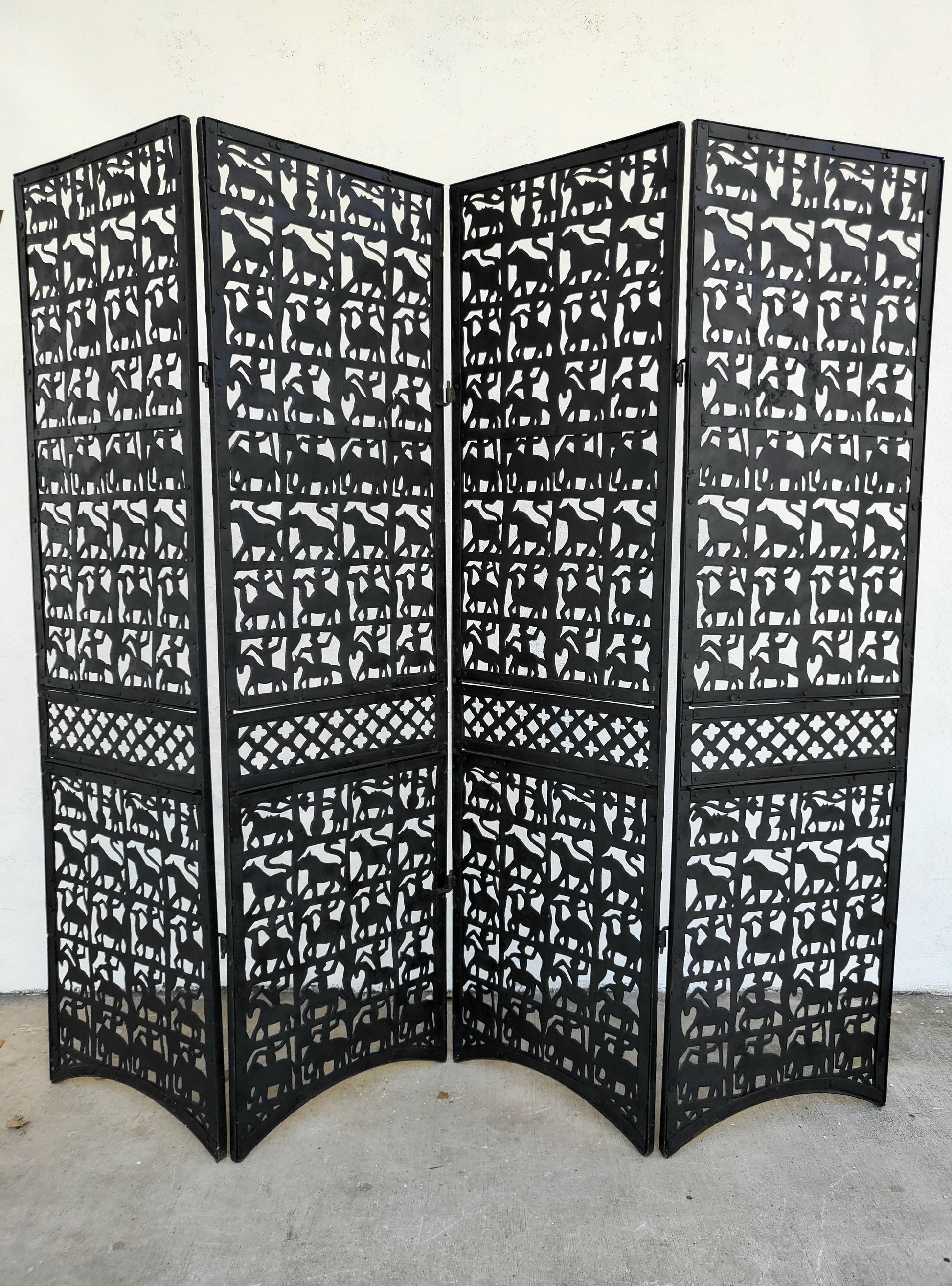 Four-panel black metal folding room divider with hand-cut animal silhouettes and lattice detailing, standing outdoors against a white wall.