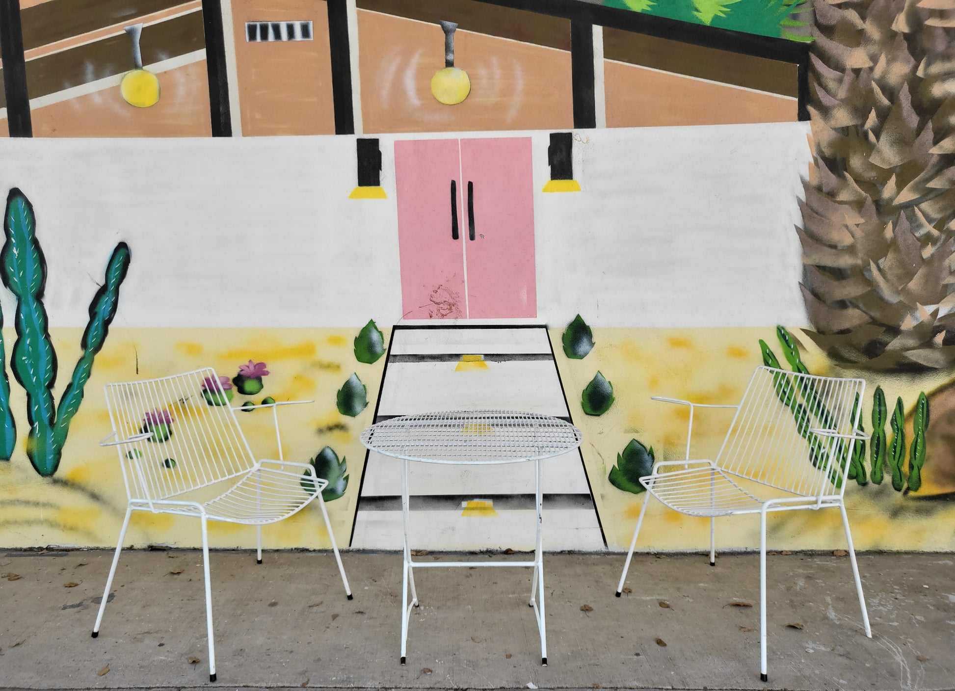 White mid-century Breotex outdoor setting with two wire chairs and a round table, set against a mural wall.