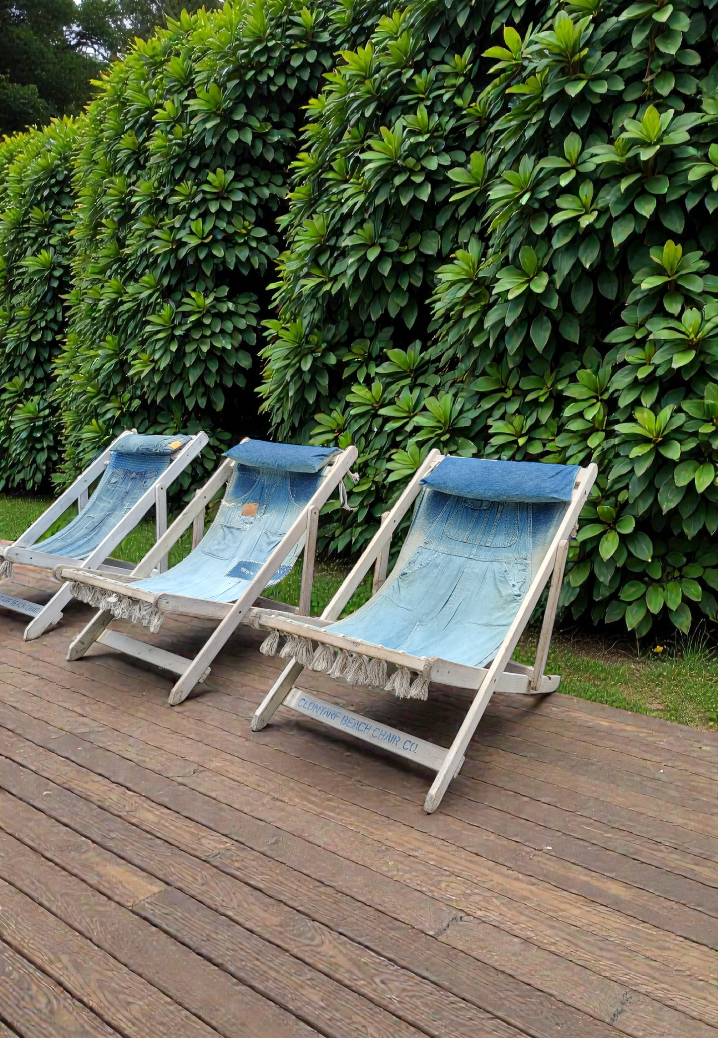 Set of three folding deck chairs with white timber frames and patchwork blue denim sling seats made from upcycled jeans, featuring pockets, tassel trim and soft headrest cushions in a coastal outdoor setting.